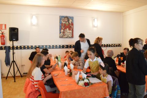 Bambini e adulti seduti a un tavolo in una mensa o sala da pranzo, apparecchiato per un pasto con tovaglie arancioni e decorazioni floreali.