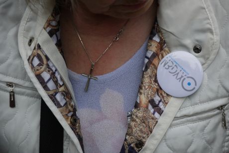 Donna con giacca chiara e spilletta sopra il petto con il logo oxygen, indossa una collana con croce.