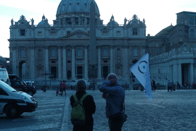Due persone in piedi di spalle in una piazza davanti a una grande basilica con facciata decorata, una di esse tiene una bandiera bianca con il logo oxygen